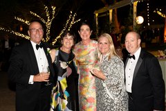 Three ladies and two men dressed in black tie attire at an outdoor evening event, string lighting in the background