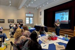 Participants seated at round tables listening to a woman in front of a screen speaking with a microphone