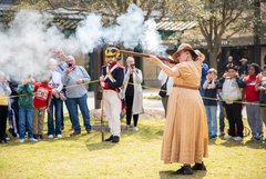 Woman in 1830s period attire firing a musket in front of a crowd of guests