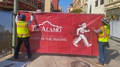 Two construction workers installing red fence wrap with Alamo logo and image of man crossing street dressed like David Crockett