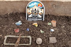 Alamo Archaeology sticker next to a group of artifacts on a dirt bed found from an excavation unit