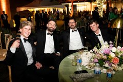 Four men in tuxedos, one wearing a coonskin cap, seated at a table with a sage linen and floral centerpiece at an evening outdoor event