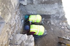 Two archaeologists wearing yellow protective vests digging inside an excavation unit