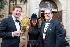 Woman in a black cowboy hat and a sleeveless black top in between two men wearing black tuxedos