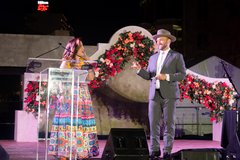 Woman in bright floral dress with man in a gray suit and hat MCing on stage at an event