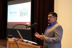Man standing at a podium speaking with a screen to the side