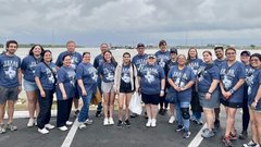 Group of people in navy blue T-shirts standing outside on a paved area in front of a lake