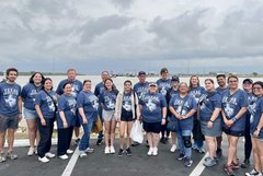 Group of people in navy blue T-shirts standing outside on a paved area in front of a lake