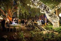 String lights hanging from trees and oak wrapped in string lights at an outdoor event in Alamo Gardens with tables and a demonstration cannon