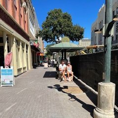 Two people seated under a canopy on a sidewalk next to a fence with black wrap
