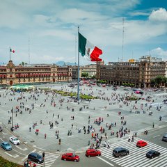 Mexican flag in city center with people in the square surrounded by buildings on two sides