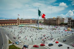 Mexican flag in city center with people in the square surrounded by buildings on two sides