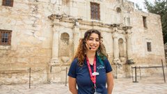 Woman with long, brown curly hair wearing a navy Alamo uniform shirt in front of Alamo Church