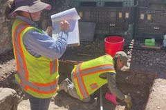 Archaeologist working in excavation area while another archaeologist takes notes
