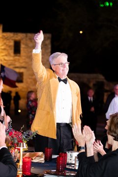 Lt. Governor Dan Patrick in a yellow jacket with his thumb up at a table in front of Alamo Church