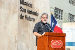 Man speaking at a podium next to the Texas flag in front of gate that says Mission San Antonio de Valero