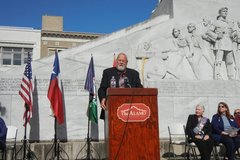 Man speaking at a podium in front of the Alamo Cenotaph