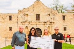 Four individuals holding a large check in front of Alamo Church