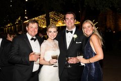 Bride, groom, and another couple smiling at an outdoor event at night with string lighting in the background