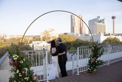 Bride and groom kiss under floral arch on terrace with view of Alamo Plaza behind them