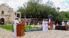 Navy officer behind a podium in Alamo Plaza with other soldiers in front of the grass for a ceremony