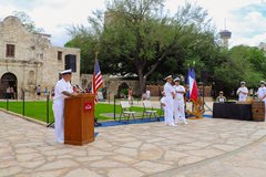 Navy officer behind a podium in Alamo Plaza with other soldiers in front of the grass for a ceremony