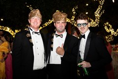 Three men in tuxedos, two with coonskin caps and one with glasses, at an evening outdoor event with string lighting in the background