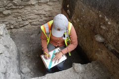 Archaeologist inside an excavation unit writing down specs holding a ruler