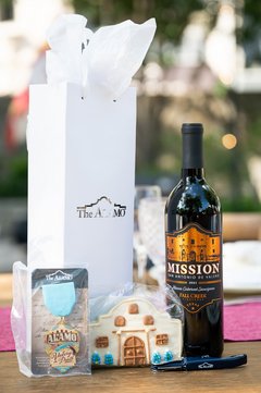 White wine bag with silver Alamo logo next to bottle of Mission wine, Fiesta medal, and Alamo shaped cookie
