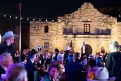 Guests seated at tables in front of Alamo Church at a formal event with string lighting