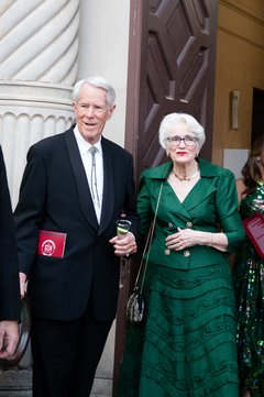 Man with white hair wering a black tuxedo with a bolo and a woman with white hair and glasses wearing an emerald green gown