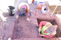 Archaeologist standing inside excavation unit with another archaeologist on the outside