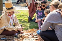 Living historian in 1830s period attire seated in a grassy area playing a card game with a young family