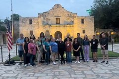 A group standing in Alamo Plaza between the US and Texas flags in front of Alamo Church