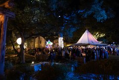 Large groups of guests under tent and throughout Alamo Gardens at night with lighted trees and installations in the background