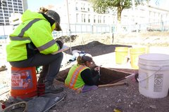 Archaeologist with yellow vest inside excavation unit, with 3 plastic buckets at the perimeter