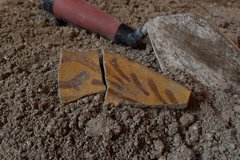 Two pieces of lead glazed rim sherds on a dirt surface next to a trowel tool