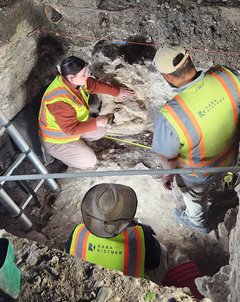 Three archaeologists wearing yellow protective vests inside an excavation unit