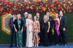 Five couples in black tie attire in front of a floral step and repeat backdrop
