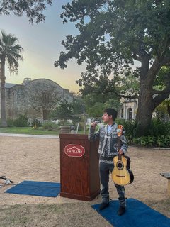 Singer holding a microphone and a guitar standing next to a podium with the Alamo logo