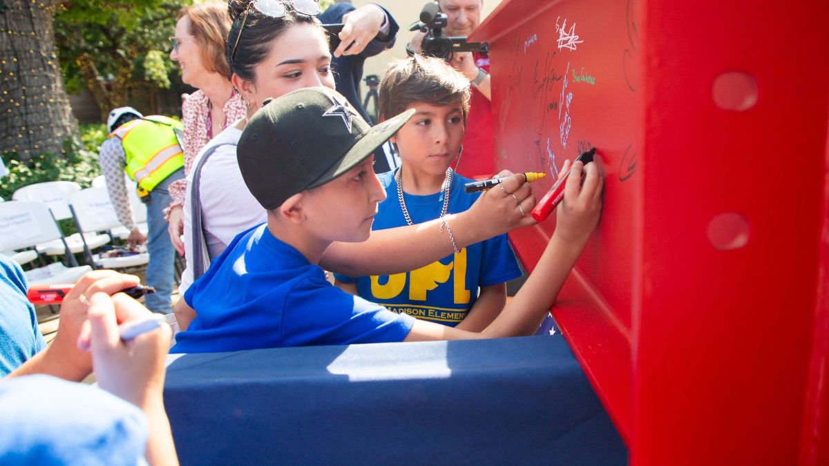 Child in a blue shirt and baseball cap signing a red metal beam