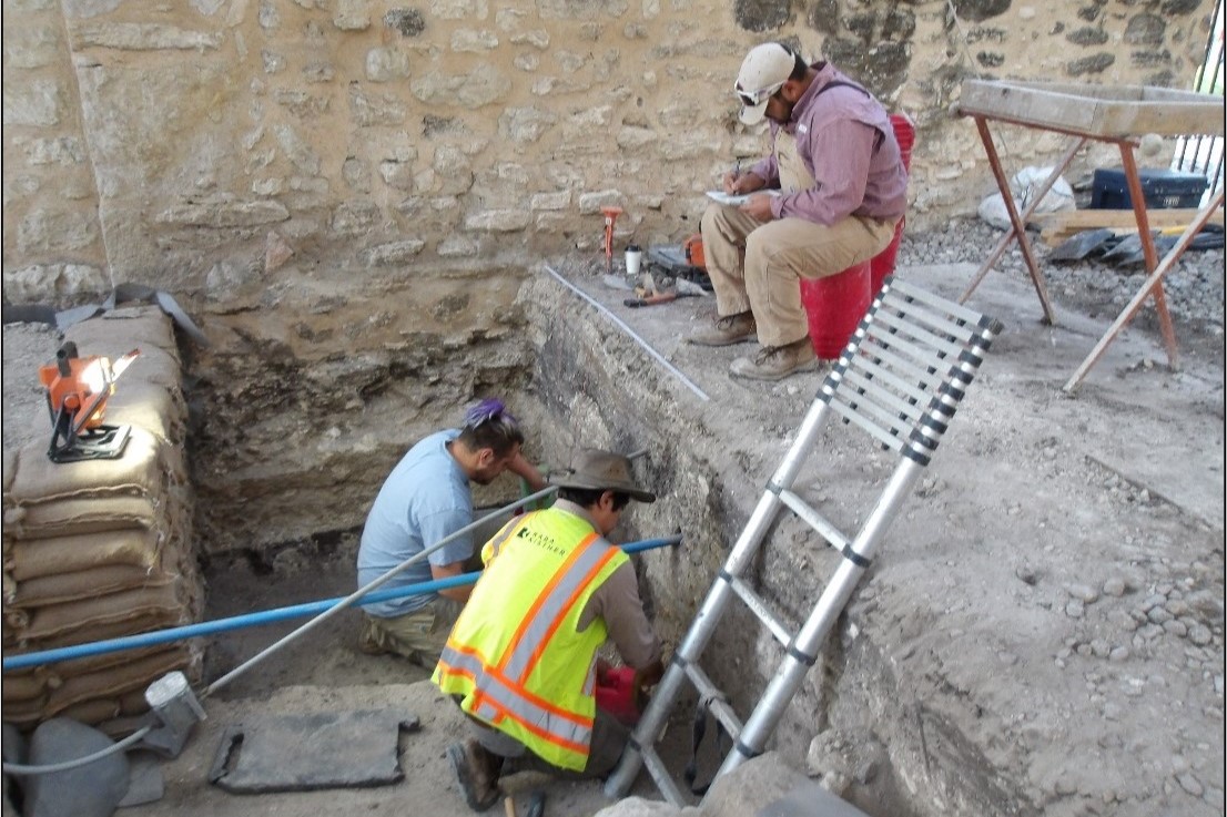 Two archaeologists inside excavation unit and one at the top with a ladder for exit
