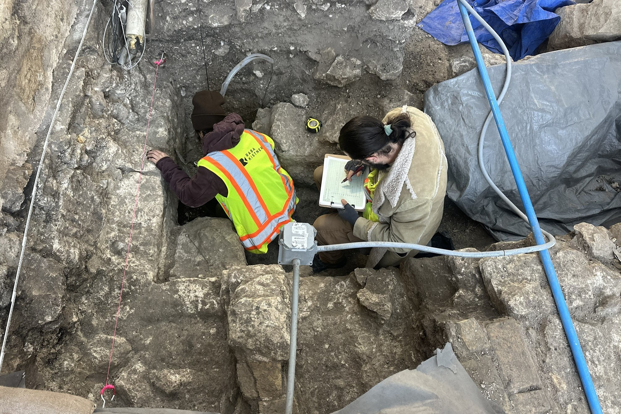 Two archaeologists inside an excavation unit facing west