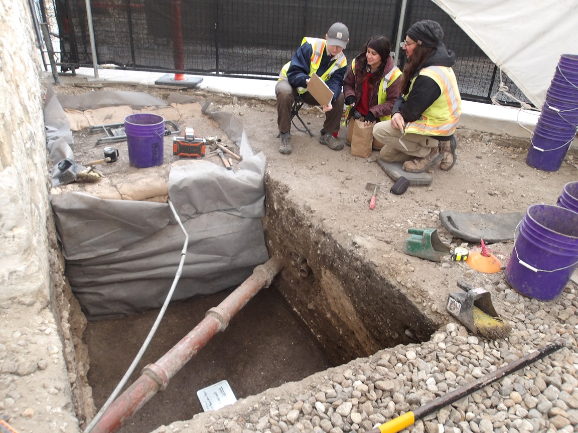 Archaeologists looking at paperwork, kneeling next to a excavation unit