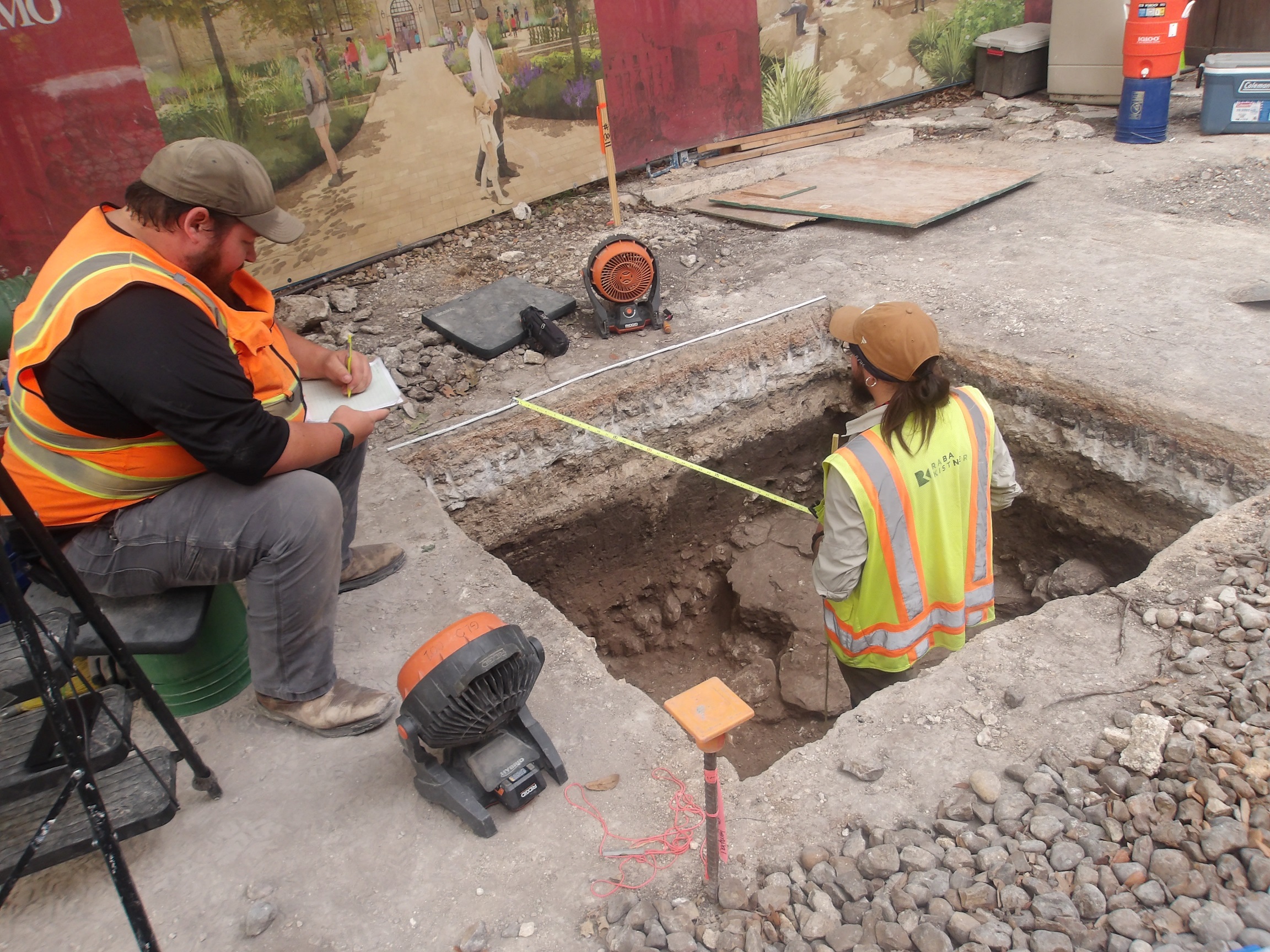 Archaeologist standing inside an excavation unit with measuring tape while another writes down the measurements