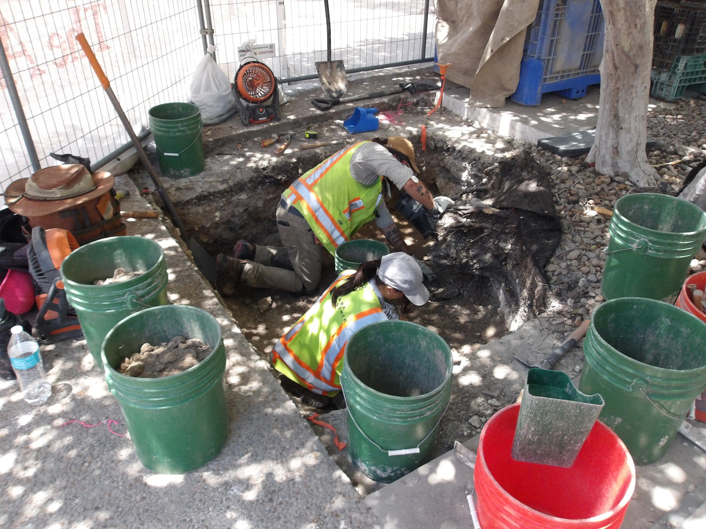 Two archaeologists working inside an excavation unit surrounded by several green buckets and one red bucket