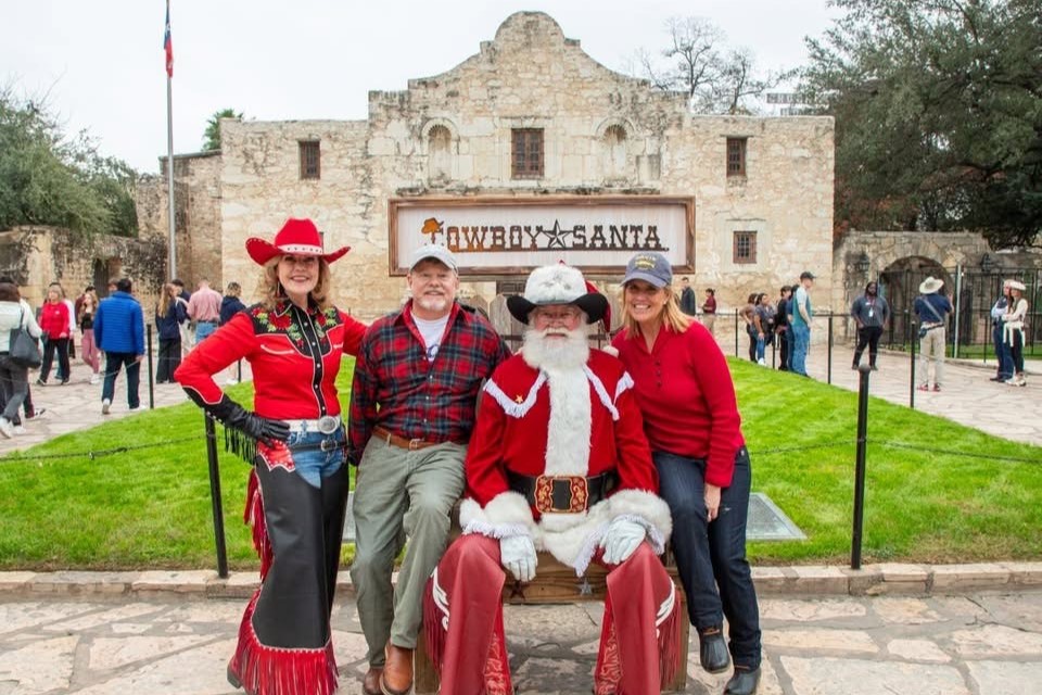 Santa wearing cowboy hat sitting in front of Alamo Church with some festive helpers